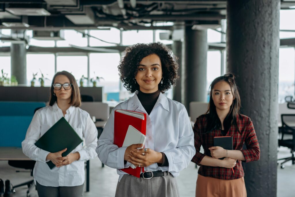 Three professional women standing confidently in a modern office environment, showcasing diversity.