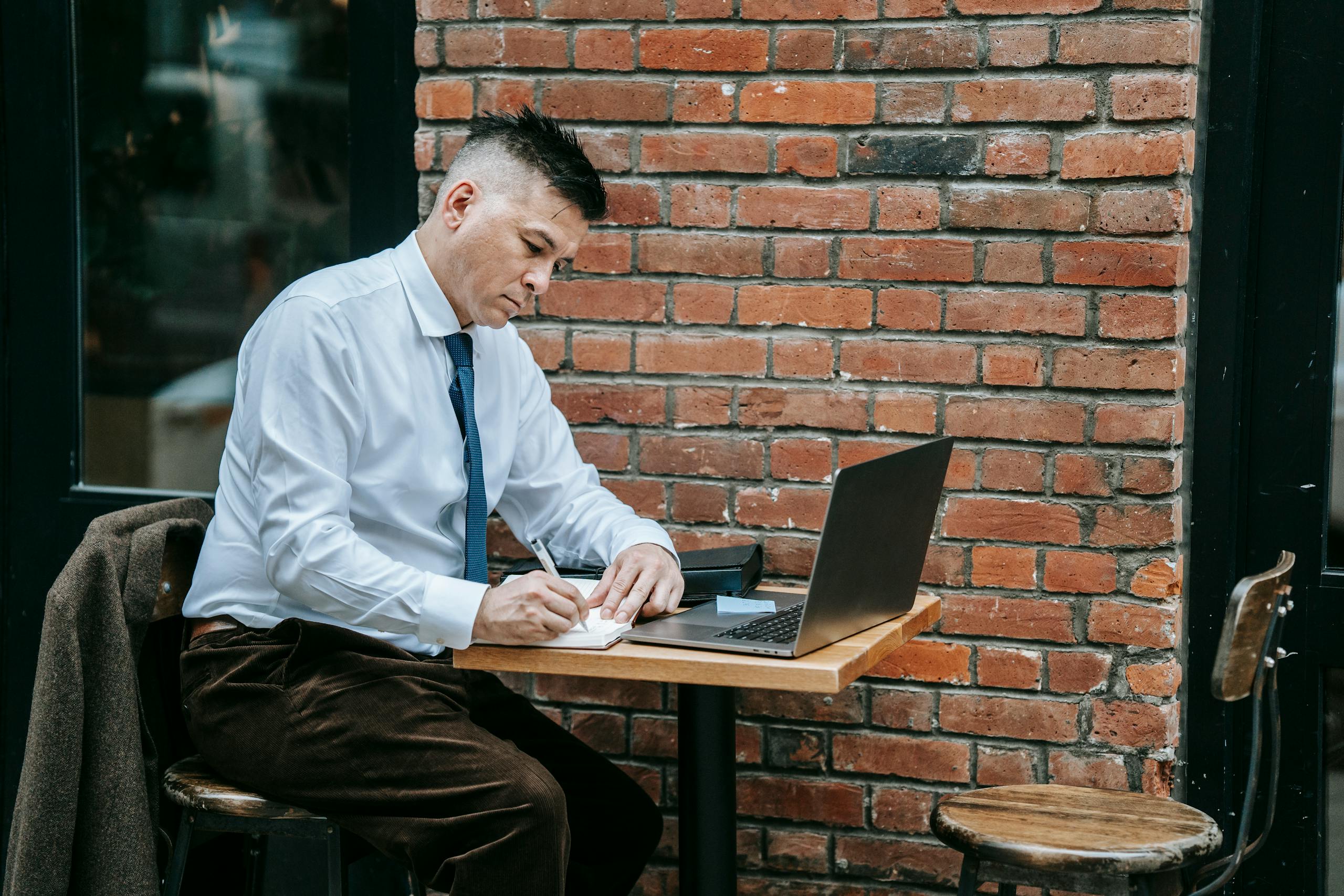Adult man in corporate attire working on laptop at a cafe table outdoors.